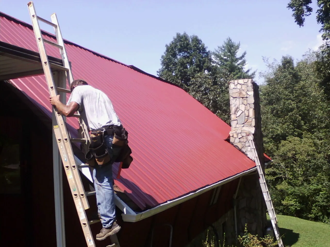 Roofer on a ladder beside a red metal roof and stone chimney — Soffit & Fascia Repair by Roofing Brothers in Charleroi